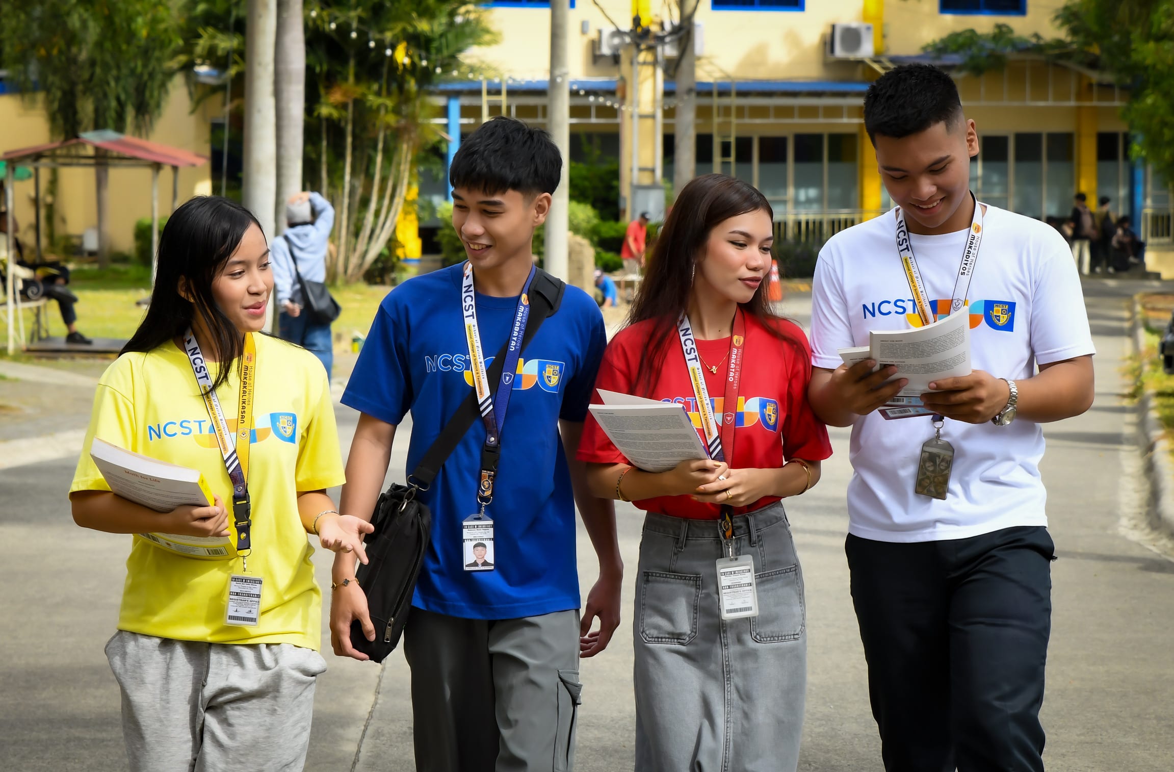 Four NCST students in programme shirts and lanyards walk together across campus while reading the documents they are carrying